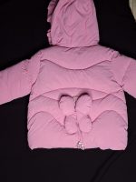 Back view of a girls' pink polyester jacket with a hood and a decorative butterfly-shaped bow on the lower back.