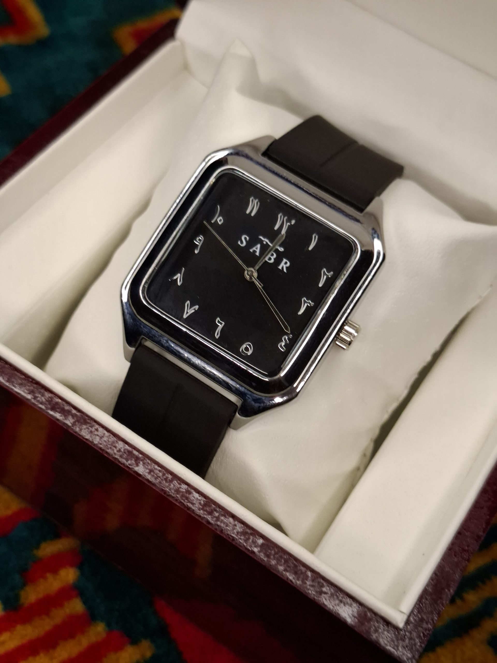 A SABR 1037B model watch with a silver square case and black dial with Arabic numerals, shown resting in its white display box.