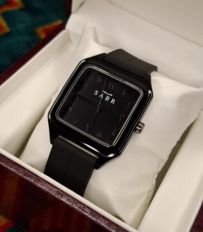 A black square-faced SABR 1037B watch with a black strap, shown from an angle inside its presentation box.