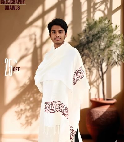 A man wearing a white Arabic calligraphy shawl with brown lettering and a fringed edge, standing indoors.