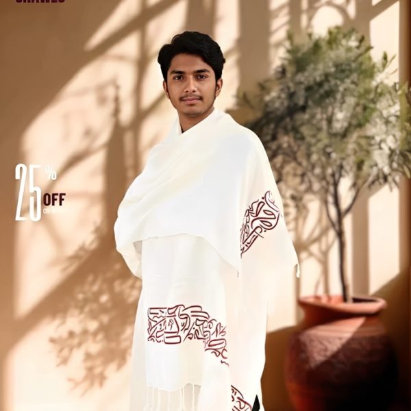 A man wearing a white Arabic calligraphy shawl with brown lettering and a fringed edge, standing indoors.
