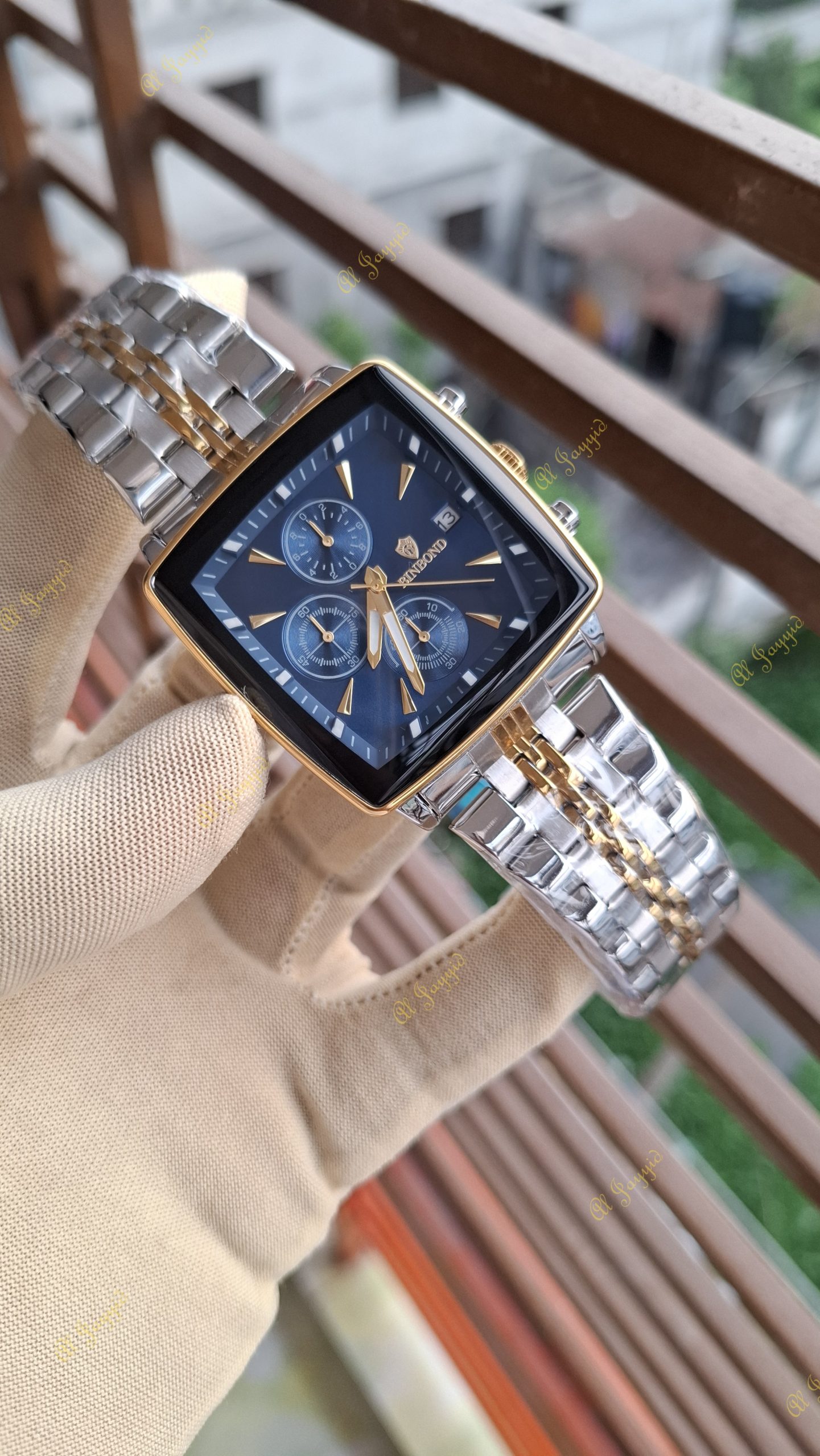 A gloved hand holds the Binbond 911 men's watch, showing its blue square dial and two-tone silver and gold bracelet.