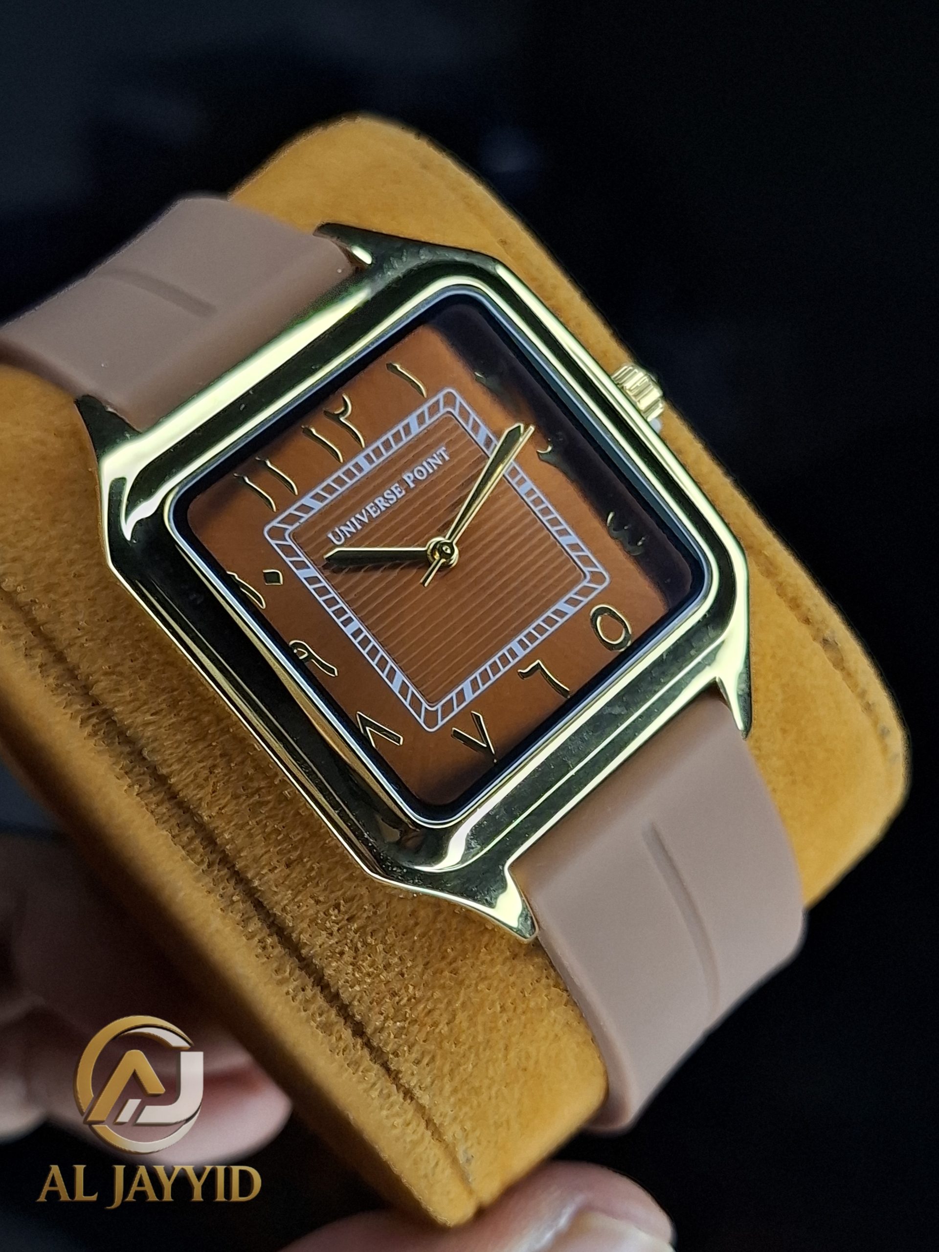 A close-up of the Universe Point Watch, featuring a square gold-tone case, a brown dial with Arabic numerals, and a beige silicone strap.