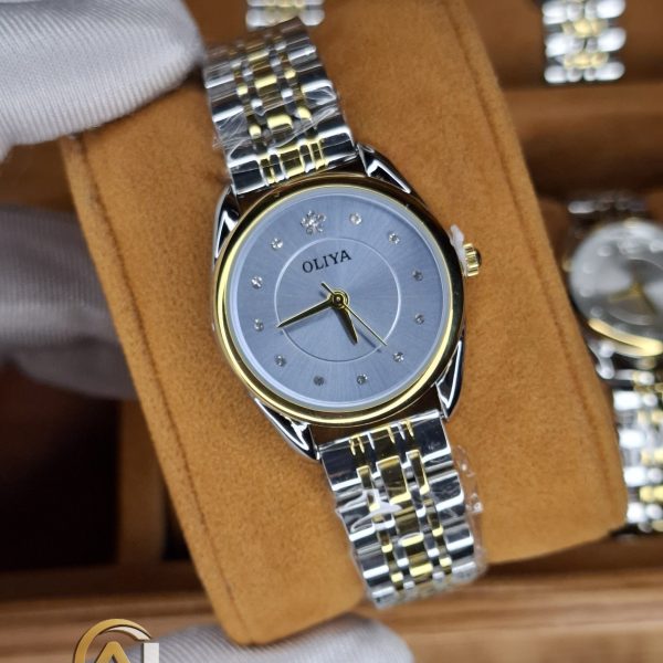 A gloved hand holding the OLIYA 6658 women's watch, which has a two-tone silver and gold band and a grey dial with crystals.