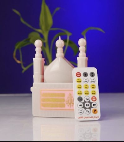 A white mosque-shaped Islamic gadget with its remote control, set against a blue background with a plant.