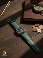 A Casio AE-1200WHL-5AV men's watch with a green strap and digital display, laid on a dark wooden desk beside a book and coins.