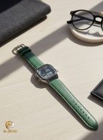 A Casio AE-1200WHL-5AV men's watch with a green leather strap and a silver case, resting on a wooden desk next to a pair of glasses.
