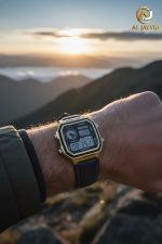 A man's wrist wearing the Casio AE-1200WHL-5AV watch with a gold case and dark strap, set against a mountain sunrise.