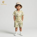 A young boy smiling and posing in a coordinated two-piece summer outfit. He is wearing a short-sleeved, button-down shirt with a Cuban collar and matching shorts, both featuring a light grey-green floral and botanical print on an ecru background. He is also wearing white socks and white sneakers, standing against a plain grey backdrop.