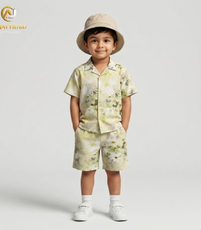 A young boy smiling and posing in a coordinated two-piece summer outfit. He is wearing a short-sleeved, button-down shirt with a Cuban collar and matching shorts, both featuring a light grey-green floral and botanical print on an ecru background. He is also wearing white socks and white sneakers, standing against a plain grey backdrop.
