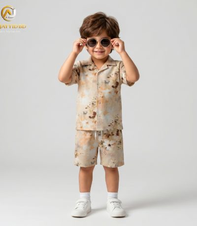 A young boy smiling and posing in a coordinated two-piece summer outfit. He is wearing a short-sleeved, button-down shirt with a Cuban collar and matching shorts, both featuring a light grey-green floral and botanical print on an ecru background. He is also wearing white socks and white sneakers, standing against a plain grey backdrop.