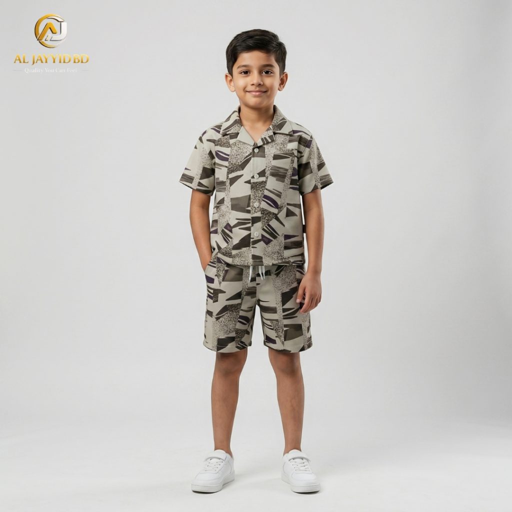 A young boy smiling and posing in a coordinated two-piece summer outfit. He is wearing a short-sleeved, button-down shirt with a Cuban collar and matching shorts, both featuring a light grey-green floral and botanical print on an ecru background. He is also wearing white socks and white sneakers, standing against a plain grey backdrop.