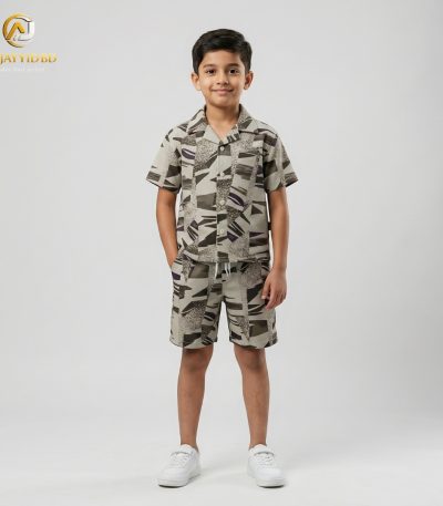 A young boy smiling and posing in a coordinated two-piece summer outfit. He is wearing a short-sleeved, button-down shirt with a Cuban collar and matching shorts, both featuring a light grey-green floral and botanical print on an ecru background. He is also wearing white socks and white sneakers, standing against a plain grey backdrop.